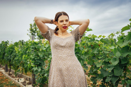 A smiling brunette corrects her hairstyle in the vineyard. A cute girl in an elegant dress makes herself a ponytail or a bun against the background of rows with grape bushes. Copy space.の写真素材