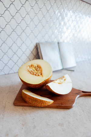 Close-up of juicy sliced melon on a wooden chopping board. An open book and a tasty fruit on the marble countertop in the kitchen. Making dessert at home. Healthy and delicious foodの写真素材