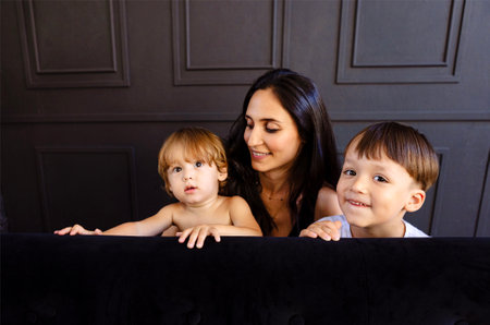 Young mother and her two little boys are playing at home. Woman in casual clothes is having fun with her cute sons in living room. Mom and her kids are laughing and hiding behind the couch. Happy familyの写真素材