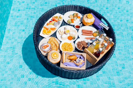 A floating breakfast tray with various dishes and drinks is located in the luxurious pool. The blue water sparkles in the sun. A place of rest and enjoyment. Lush greenery on the backgroundの写真素材