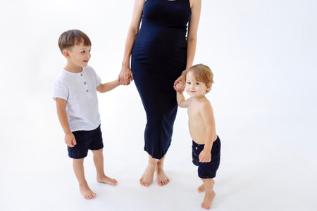 Pregnant woman holds hands with two cute children. Young mother in black dress with her kids stands barefoot on the floor. A lady expecting a baby and her smiling little boys. White isolated backgroundの写真素材