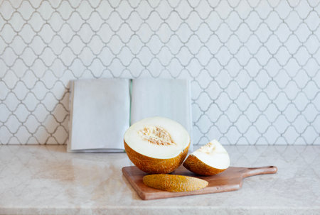 Close up of juicy sliced melon on a wooden chopping board. An open book and a tasty fruit on the marble countertop in the kitchen.の写真素材