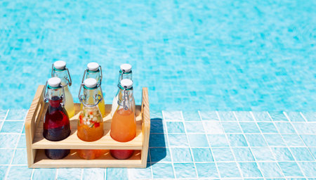 Close-up of a wooden box with glass bottles on the side of the pool. Delicious chilled multicolored drinks on a background of clear blue water. Bright natural sunlight. Rest, travel and vacationsの写真素材