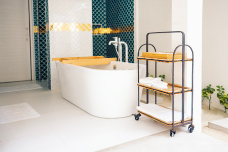 Stylish mobile shelf with clean towels in the bathroom. The modern room is decorated with blue and white tiles in the form of fish scales. A coffee serving table and bathtub with a wooden shelfの写真素材