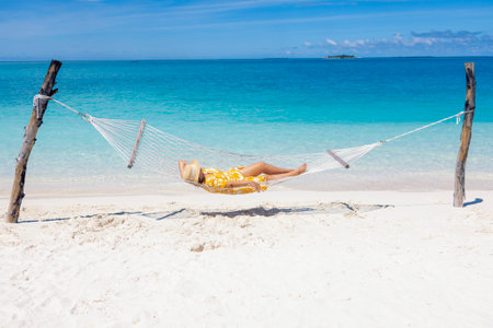 A young woman in a straw hat and a summer dress is lying in a hammock near the ocean. The lady sleeps and relaxes on the seashore. The girl is enjoying her vacation and the sea breeze on the beachの写真素材