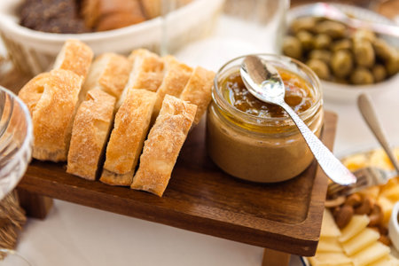 A close-up of a wooden tray with slices of fragrant fresh bread and a jar of peanut paste. Delicious snacks and a plate of olives on a white tablecloth. A hearty breakfast or lunchの写真素材