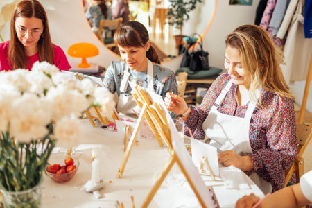 Young women in casual clothes at a painting workshop. Friends at a table with easels and blank canvases. The view from the side. Girls learn to draw in an art studio. A creative hobbyの写真素材
