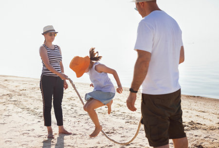 Mom and dad are holding a rope, their daughters are jumping over it. A young married couple having fun with a child by the sea. Parents and their children play and laugh on the sandy beach. A sunny day of summer vacationの写真素材