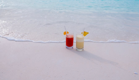 Two colorful delicious cocktails on the ocean. Glasses with cool drinks and pieces of fruit near the clear blue water on the shore. Milkshake and fresh orange juice on a clean sandy beach on vacationの写真素材