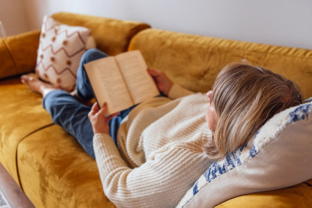 An elderly woman in casual clothes is lying on the couch and reading a novel. A mature lady holds a book and relaxes at home on the sofa in the bedroom. Stylish, modern and cozy interiorの写真素材