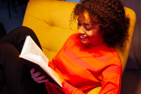 Young laughing African woman in sweater is sitting in yellow armchair and reading. Attractive black...