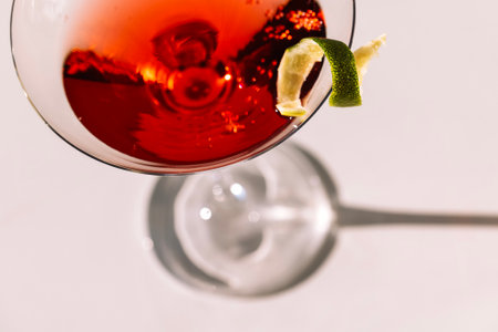 Close-up of a martini glass on a white table. An elegant goblet with a red alcoholic sparkling drink decorated with lime zest. Tasty berry cocktail on a minimalist background. Sunlightの写真素材