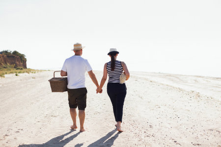 A married couple goes on a picnic by the sea. A man and his wife in casual clothes are walking on a sandy beach. A smiling woman and her husband hold hands and have fun together. A romantic meetingの写真素材