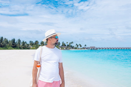 A young man stands on the ocean shore. A middle-aged male wearing sunglasses and a hat rests on a tropical island. Tourist enjoys the seascape and is relaxing on the beach on a summer vacationの写真素材