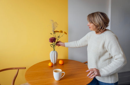 An elderly woman in a white sweater and jeans collects an autumn bouquet in a vase on a wooden round table. A mature lady decorates her house with flowers and drinks coffee. Stylish modern interiorの写真素材