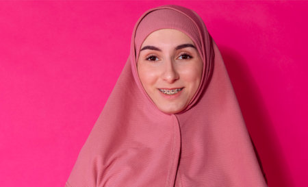 An Arab girl in a pink hijab on a pink background. A cute teen in Muslim religious clothes smiles against a bright backdrop. A female teenager with braces in an Islamic headscarf poses in a studioの写真素材