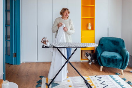 Close-up of an elderly woman ironing clothes on an ironing board. A mature lady holds an iron and smiles at home. The pensioner does household chores. Cozy and stylish interiorの写真素材