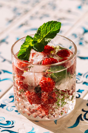 Close-up of a glass of mineral water with raspberries and fresh mint and ice cubes on a table made of white tiles with blue patterns. A cup of cool, delicious, healthy drink. Copy spaceの写真素材