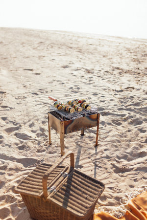 Barbecue with vegetable kebabs on the sandy beach. Zucchini, eggplant and tomatoes are grilled over a fire. A delicious healthy snack. Wicker picnic basket by the sea. Summer vacationの写真素材