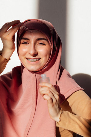 A young girl in a pink hijab applies a transparent moisturizing serum to her face. A teen in a Muslim headscarf smiles and holds a bottle of essence. A female with an dropper and a cosmetic productの写真素材