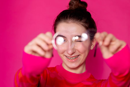 A smiling girl holds out her glasses and winks at a pink background. Cute teen with braces holds glasses on a bright background. A female teenager in a casual striped sweater with glasses in her handsの写真素材