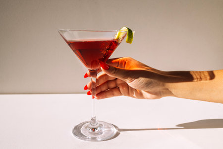 Close-up of a martini glass and a female hand with a red manicure. A woman holds a goblet with a alcoholic drink decorated with lime zest. Tasty berry cocktail on a minimalist background. Sunlightの写真素材