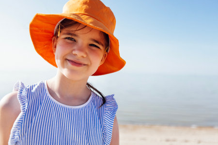 Close-up portrait of a little girl in an orange panama hat smiling by the sea. A cute child in casual clothes is relaxing on the beach. Sweet baby is enjoying summer vacationの写真素材