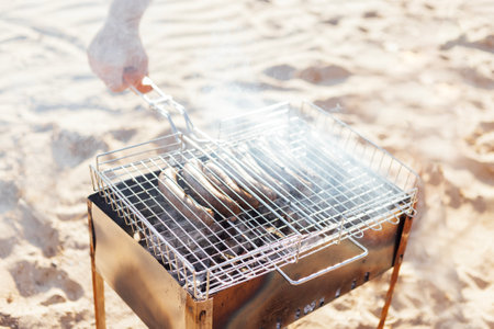 Close-up of a man hand holding a hand grill with fried sausages. Barbecue grill on the sandy beach. A man roasts sausages on an open fire. Picnic on the seashore.の写真素材