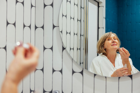 An elderly woman in a white terry dressing gown applies a serum to her face. A mature lady takes care of her skin and looks at herself in a round mirror in the bathroom at home. Stylish interiorの写真素材