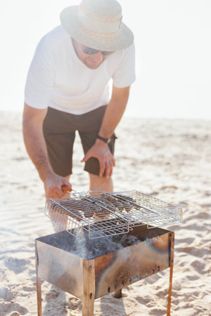 A young man in a summer casual outfit is having a barbecue on the sandy beach. A male in sunglasses and a straw hat is frying sausages on the seashore. A man cooks grilled meat over an open fireの写真素材