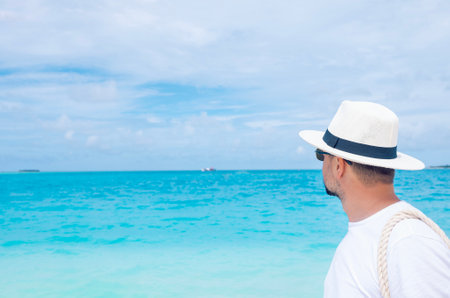 A young man stands on the ocean shore. A middle-aged male wearing sunglasses and a hat rests on a tropical island. Tourist enjoys the seascape and is relaxing on the beach on a summer vacationの写真素材