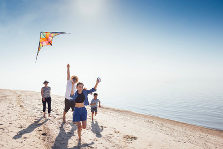 A happy family is relaxing during the summer holidays. Children, their parents fly a kite on the riverbank. A married couple with their kids are running along the sandy beach and having fun togetherの写真素材