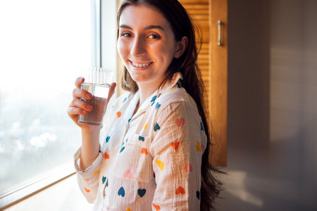 Close-up of a charming girl in cute pajamas standing by the window and drinking water. A young student has woken up and is holding a glass of water in her bedroom at home. Good morning teenの写真素材