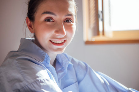 Close-up portrait of a smiling girl in a blue shirt. A young woman laughs and sits next to the window. A happy teenage girl in casual clothes in her modern cozy roomの写真素材