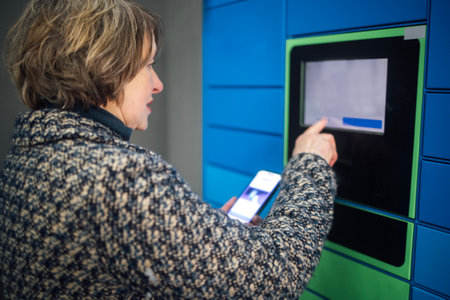 An elderly woman taps the display to receive a parcel. A mature lady in casual clothes enters data on a parcel automat. The pensioner holds the phone and dials the code to receive the purchaseの写真素材