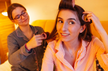 Close up of a smiling girl in pajamas with curlers. Friends laugh and put their hair in curlers before going to bed. Teenagers have fun together and do their hairstylesの写真素材