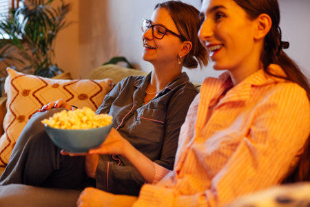 Girls in pajamas are sitting on the couch watching a movie. Friends are smiling and eating popcorn. Students are having fun together at home and laughing at a comedy. Happy sisters are watching TVの写真素材