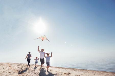 A happy family is relaxing during the summer holidays. Children, their parents fly a kite on the riverbank. A married couple with their kids are running along the sandy beach and having fun togetherの写真素材