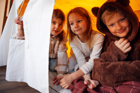 Small smiling children play in a tent at home. Friends are having fun and sitting under the table on a mattress in the living room. Girls and a boy laugh together in a bedroom in a children playhouseの写真素材