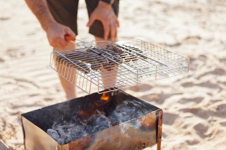 A young man in a summer casual outfit is having a barbecue on the sandy beach. A male in sunglasses and a straw hat is frying sausages on the seashore. A man cooks grilled meat over an open fireの写真素材