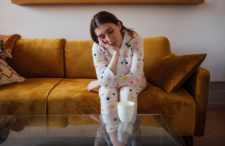 Close-up of a funny girl in cute printed pajamas sitting on the couch and yawning. A teenager is dozing while sitting on the sofa. A sleepless girl with a cup of coffee at homeの写真素材