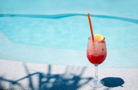 A delicious berry cocktail in an elegant glass on the side of the pool. A cup of cool summer drink with a slice of orange on a background of blue water. Tasty juice with ice in bright sunlightの写真素材