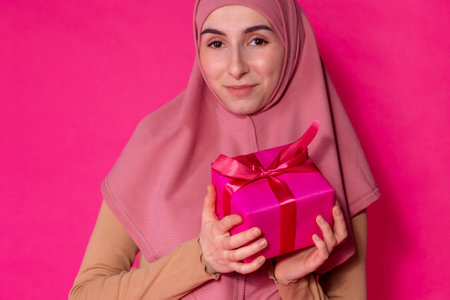 An Arab female teen in a pink hijab holding a gift. A girl in Muslim religious clothes smiles against a bright background. A teenager with braces wearing an Islamic women headscarf with a present boxの写真素材