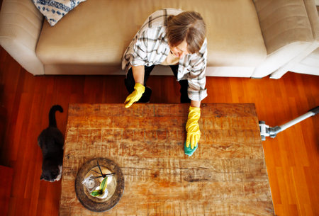 An elderly woman in casual clothes washes a wooden coffee table in the living room. A mature lady is...