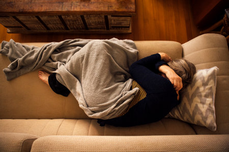 An elderly woman lies covered with a blanket and suffers from a headache at home. A mature lady sleeps on the couch and hugs herself by the shoulders. Top view. A sick pensioner is curled up and lying on the sofaの写真素材