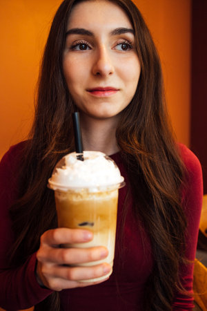 A young girl with long dark hair and casual clothes with a glass of drink. A teenager holds a delicious cocktail with whipped cream and a straw. A thoughtful girl in a cafe with a tasty milkshakeの写真素材