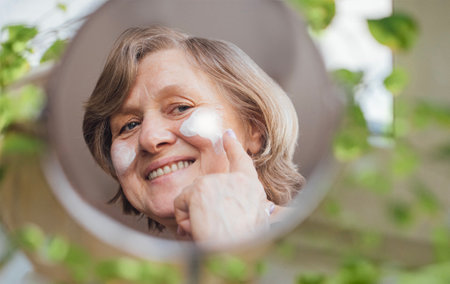 An elderly woman applies cosmetics product to her face. The reflection of a pensioner with cream in a mirror. A mature lady smears her skin with a moisturizer. Skin care in adulthoodの写真素材