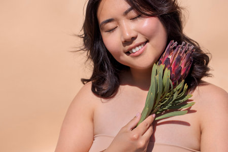 Smiling young woman with dark hair holds a colorful protea flower, radiating happiness and warmth against a soft background, creating a serene and inviting atmosphereの写真素材