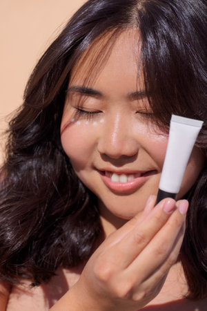 Smiling female with dark hair holds skincare tube, radiating joy and confidence in a bright, natural...