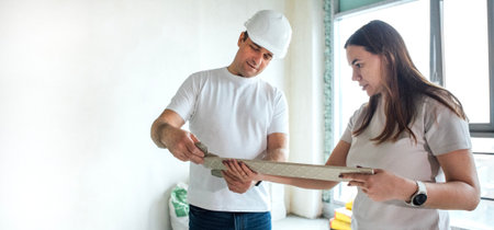 A young man in a construction helmet shows a woman a tile sample or building material. A girl examines a finishing element attentively. Repair of a house or arrangement of an apartment interior.の写真素材